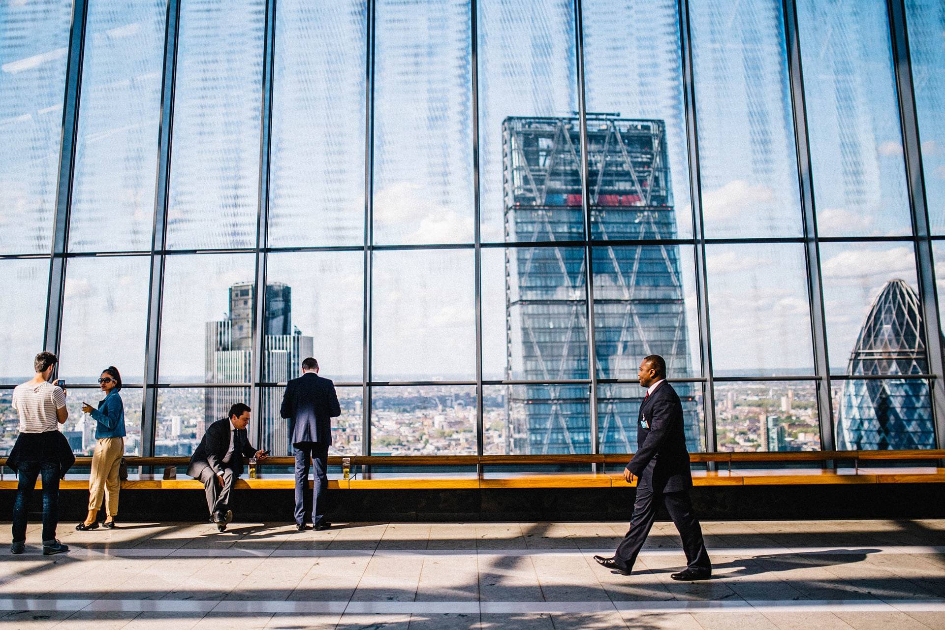 Auf diesem Bild sieht man einen Teil eines grossen modernen Firmengebäudes, welches komplett aus Glas besteht. In diesem Glas spiegelt sich eine Stadt mit vielen Häusern und drei Hochhäusern im Vordergrund. Ebenfalls ist der hellblaue mit weissen Wolken durchzogene Himmel im Glas zu erkennen. Vor dem Firmengebäude steht eine lange Holzbank. Auf dieser Sitzt ein Mann im Anzug und schaut auf sein Handy. Vor ihm steht ein Mann mit dem Rücken zu uns. Links vom Bild sieht man wie ein Mann eine Frau Fotografiert. Von rechts her läuft ein Mann im Anzug durch das Bild.