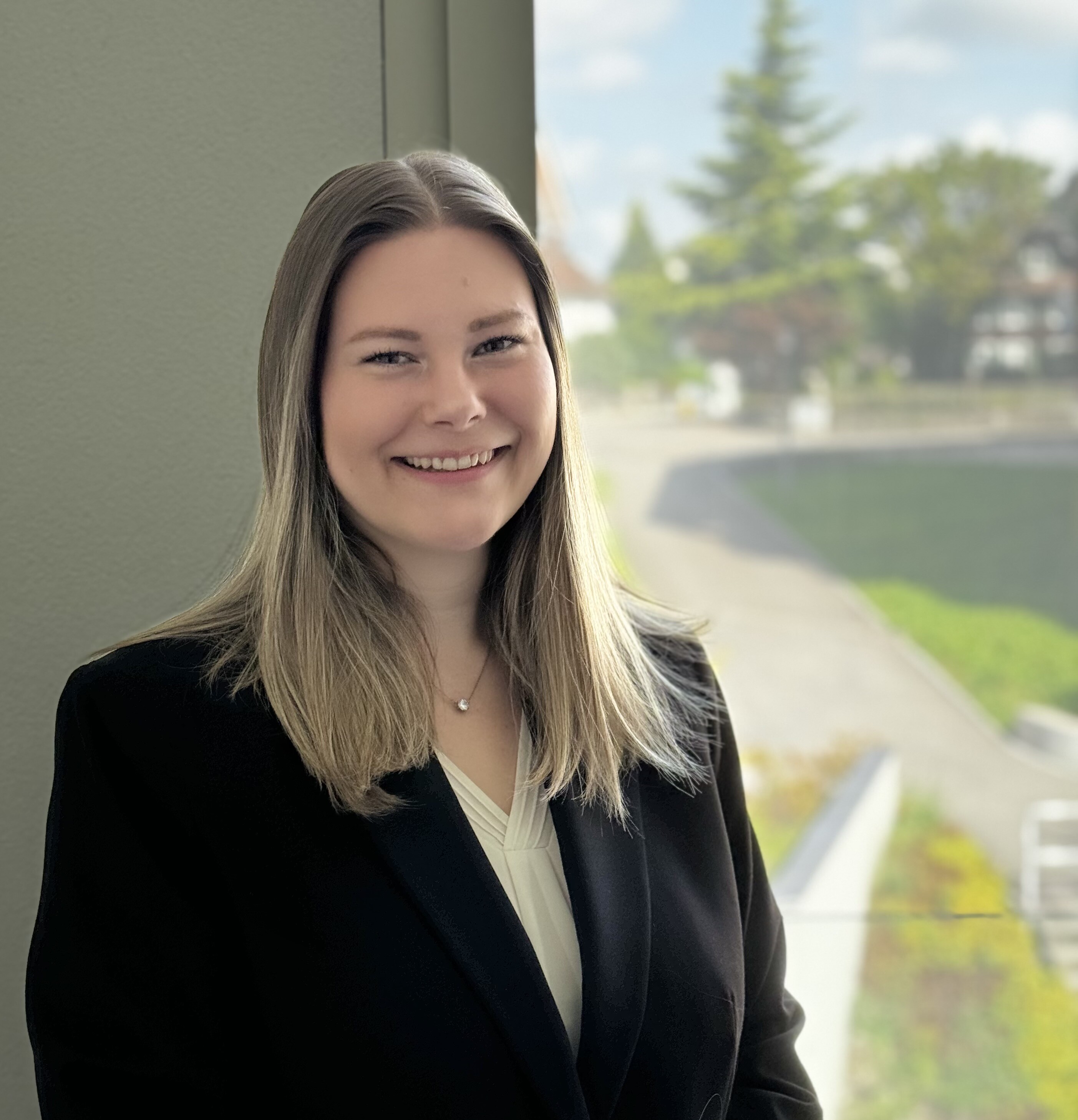 Auf dem Bild ist die Inhaberin Lea Gneiss zu sehen. Sie trägt einen schwarzen Blazer und darunter eine weisse Bluse. Sie trägt eine silberne Kette und hat mittellange dunkelblonde Haare. Im Hintergrund sieht man einen Ausschnitt von einem Fenster. In diesem Fenster ist verschwommen eine kleine Strasse, eine grüne Wiese und grüne Bäume zu sehen. Der Himmel ist blau mit wenigen weissen Wolken.